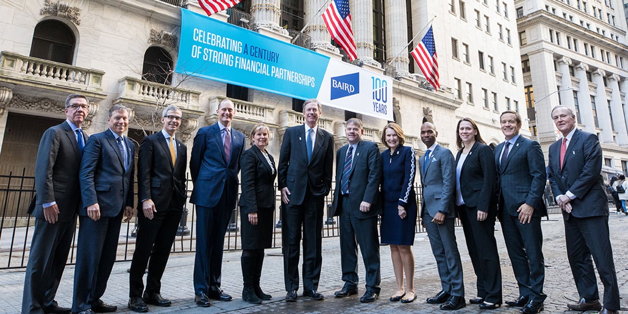  Baird leadership standing in front of the New York Stock Exchange with a banner celebrating Baird 's 100th anniversary in the background.